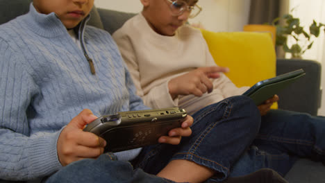 Two-Young-Boys-Sitting-On-Sofa-At-Home-Playing-Games-Or-Streaming-Onto-Digital-Tablet-And-Handheld-Gaming-Device-4